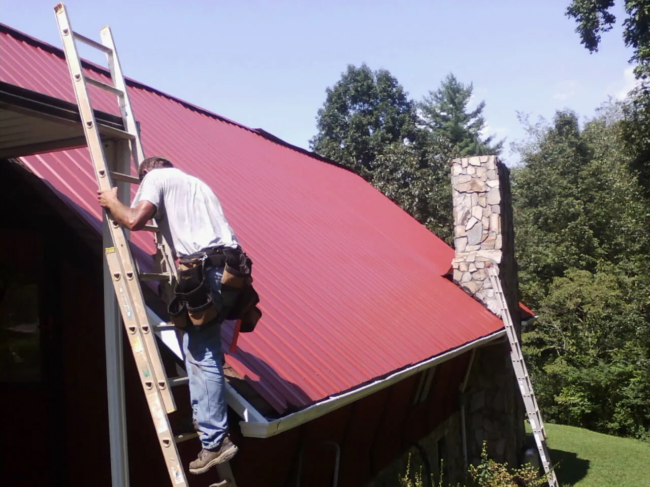 Roofer on a ladder beside a red metal roof and stone chimney — Soffit & Fascia Repair by Roofing Brothers in Sag Harbor